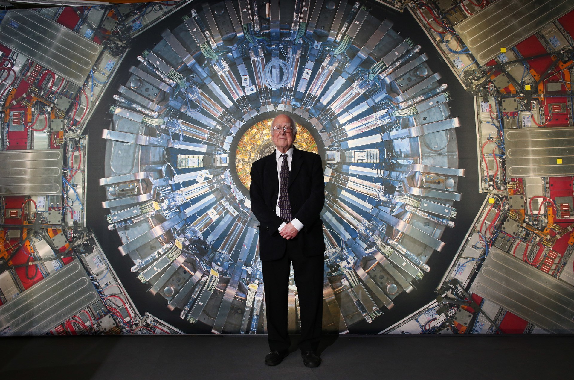 Professor Peter Higgs stands in front of a photograph of the Large Hadron Collider.