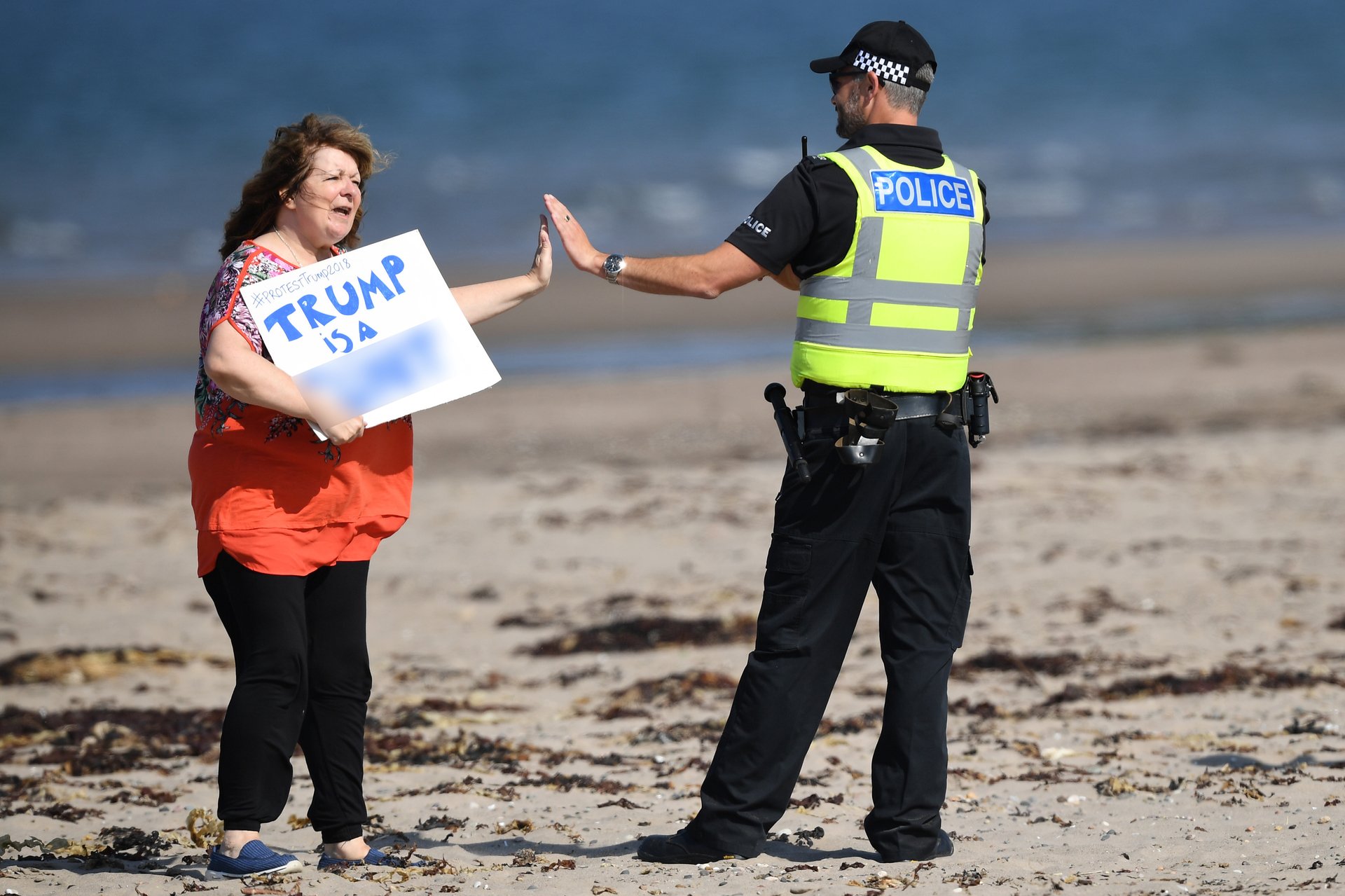 Janey Godley gives a high-five to a police officer as she holds a sign stating 'Trump is a c***' on the beach outside Trump Turnberry Resort during the US President's visit to the United Kingdom on July 14, 2018.